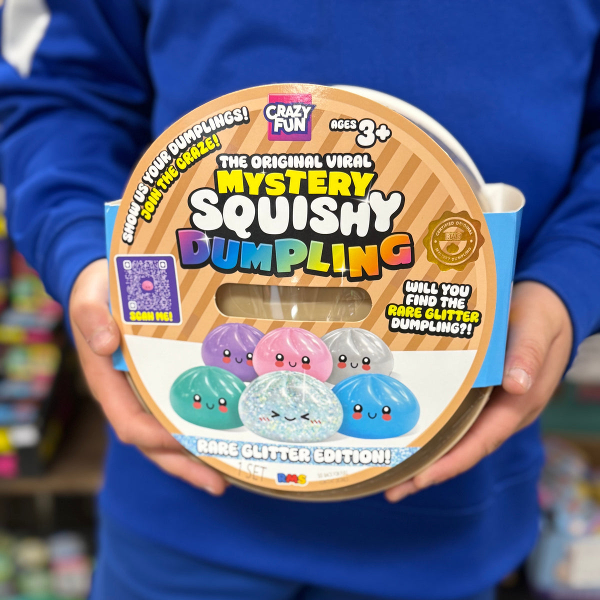 A person in a blue uniform holds a round package of 'The Original Viral Mystery Squishy Dumpling' by Crayshay Fun. The packaging features a tan and brown striped background with colorful cartoon dumpling graphics in purple, pink, blue, and green.