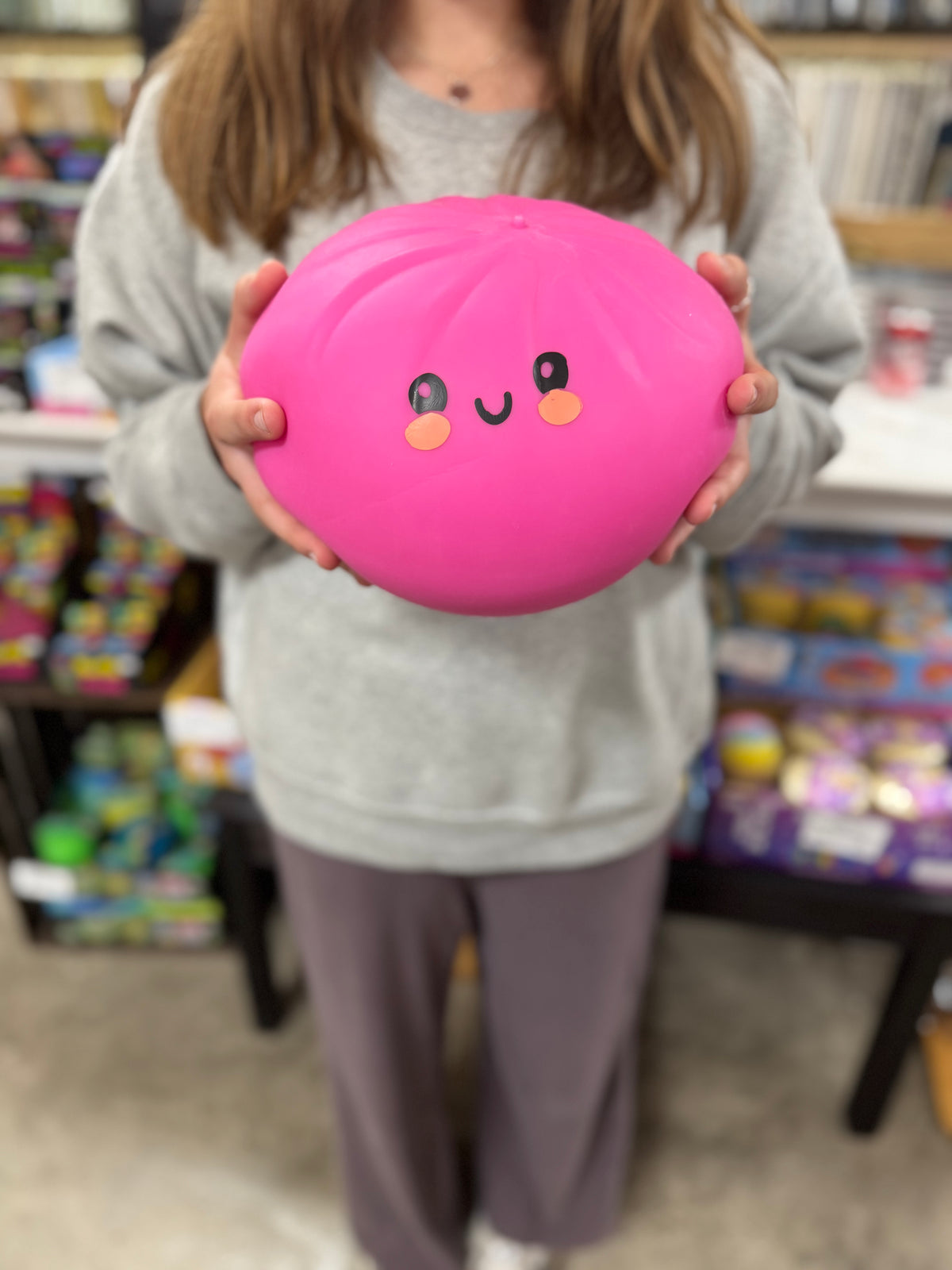 A person in a gray sweatshirt and purple sweatpants holds a bright pink, rounded squishy toy with a cute face in a retail store setting.