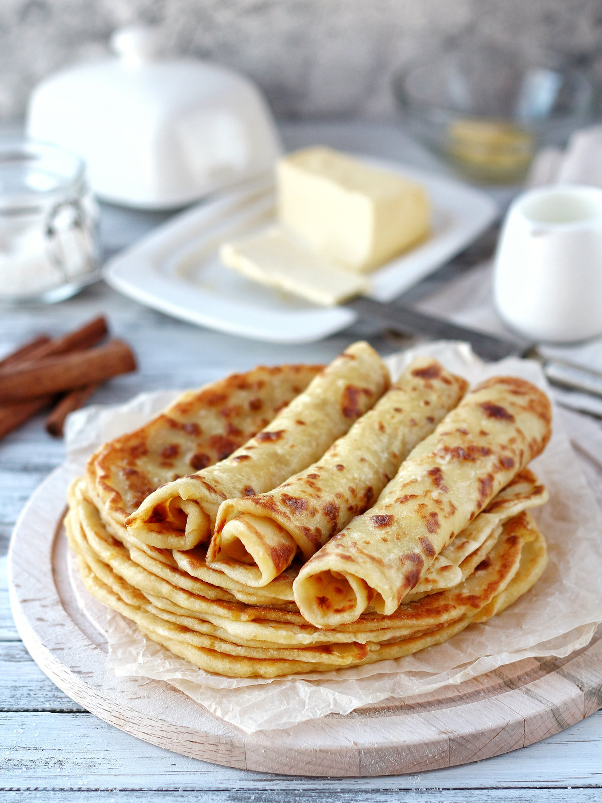 A stack of golden-brown rolled lefse on parchment paper, served on a round wooden board with cinnamon sticks, butter, and small white containers in the background.