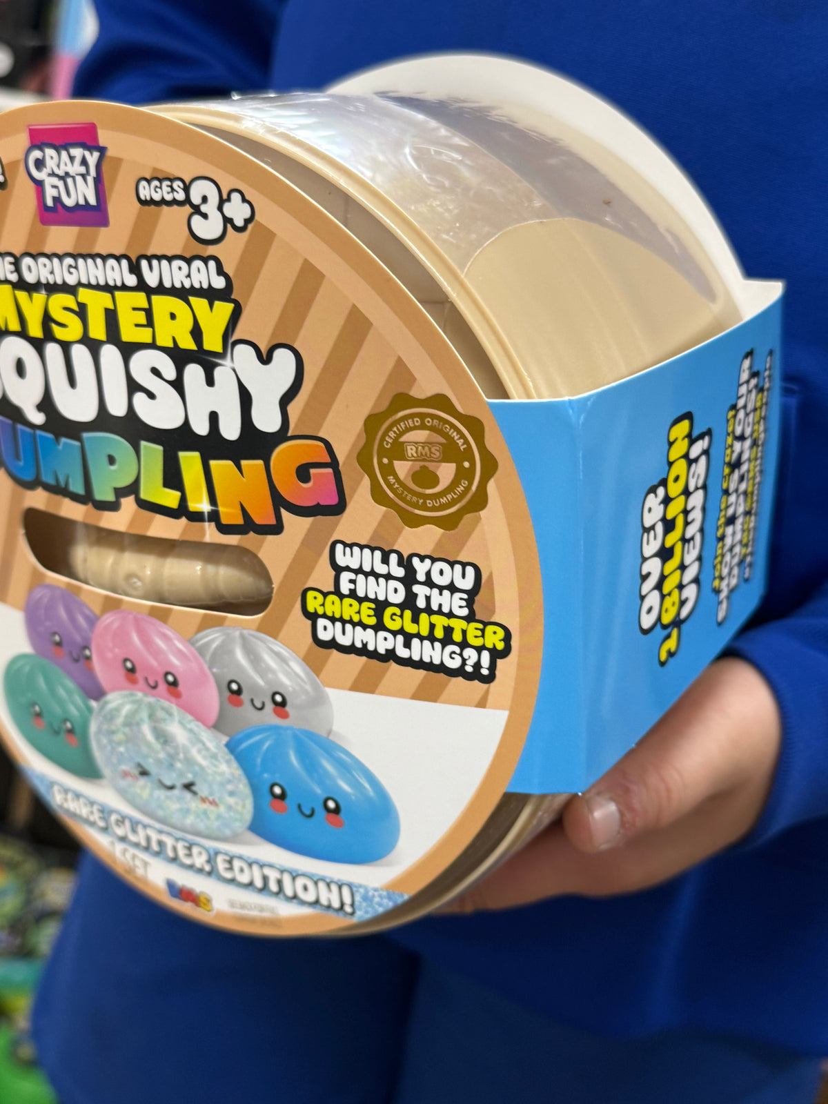 A person in a blue shirt holds a round plastic tub of Crazy Fun Mystery Quishing Glitter Edition toy capsules. The packaging features a brown and beige striped design with colorful cartoon quishing graphics and text highlighting the 'Rare Glitter Edition'.