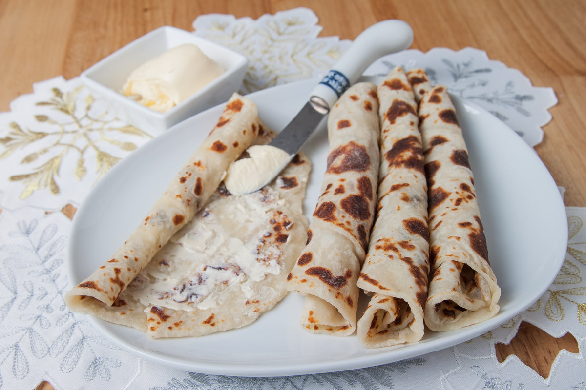 Four rolled lefse with toasted spots are served on a white oval plate, with a small dish of butter and a butter knife on a patterned white placemat over a wooden table.