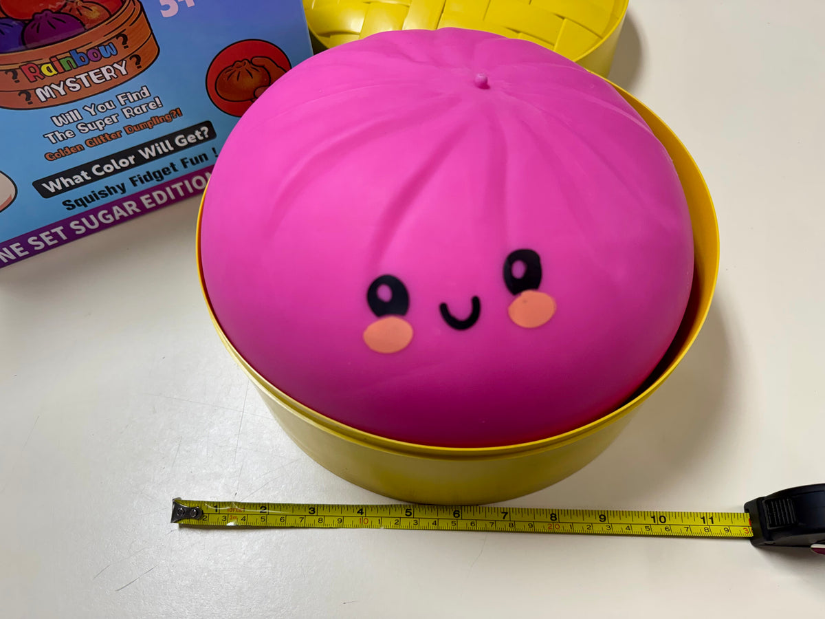 A bright pink round squishy toy with a cute face sits inside a yellow container on a white surface, with a measuring tape below and part of a product box visible in the background.