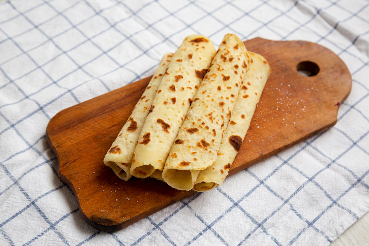 Three rolled lefse with toasted spots, stacked on a wooden cutting board over a blue-and-white checkered cloth.