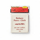 A packaged Bethany Housewares pastry cloth in clear plastic wrapping with a red and white header card, placed on a gray textured surface. The cloth itself is off-white with red printed text listing food types it resists, including pie crust, cookies, pizza, and lefse.