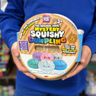 A person in a blue uniform holds a round package of 'The Original Viral Mystery Squishy Dumpling' by Crayshay Fun. The packaging features a tan and brown striped background with colorful cartoon dumpling graphics in purple, pink, blue, and green.