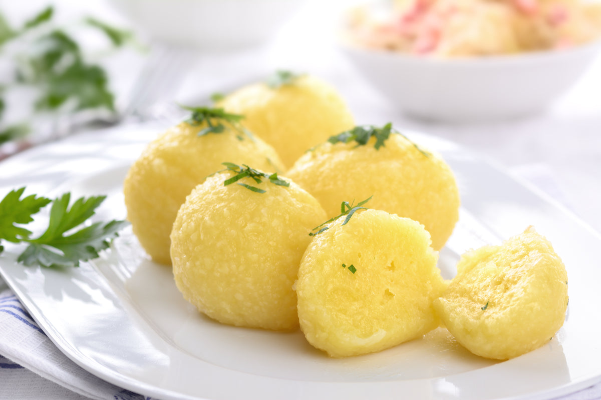 Five golden-yellow, soft-textured dumplings garnished with chopped herb, served on a white plate with parsley and a blurred side dish in the background.