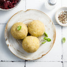 Three round golden-beige dumplings, garnished with small green herb leaves, served on a rustic beige ceramic plate. The plate is set on a white tiled surface alongside a bowl of dark red cabbage, a pepper shaker, and a bowl of peppercorns.