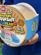 A person in a blue shirt holds a round plastic tub of Crazy Fun Mystery Quishing Glitter Edition toy capsules. The packaging features a brown and beige striped design with colorful cartoon quishing graphics and text highlighting the 'Rare Glitter Edition'.
