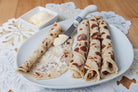 Four rolled lefse with toasted spots are served on a white oval plate, with a small dish of butter and a butter knife on a patterned white placemat over a wooden table.