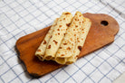 Three rolled lefse with toasted spots, stacked on a wooden cutting board over a blue-and-white checkered cloth.